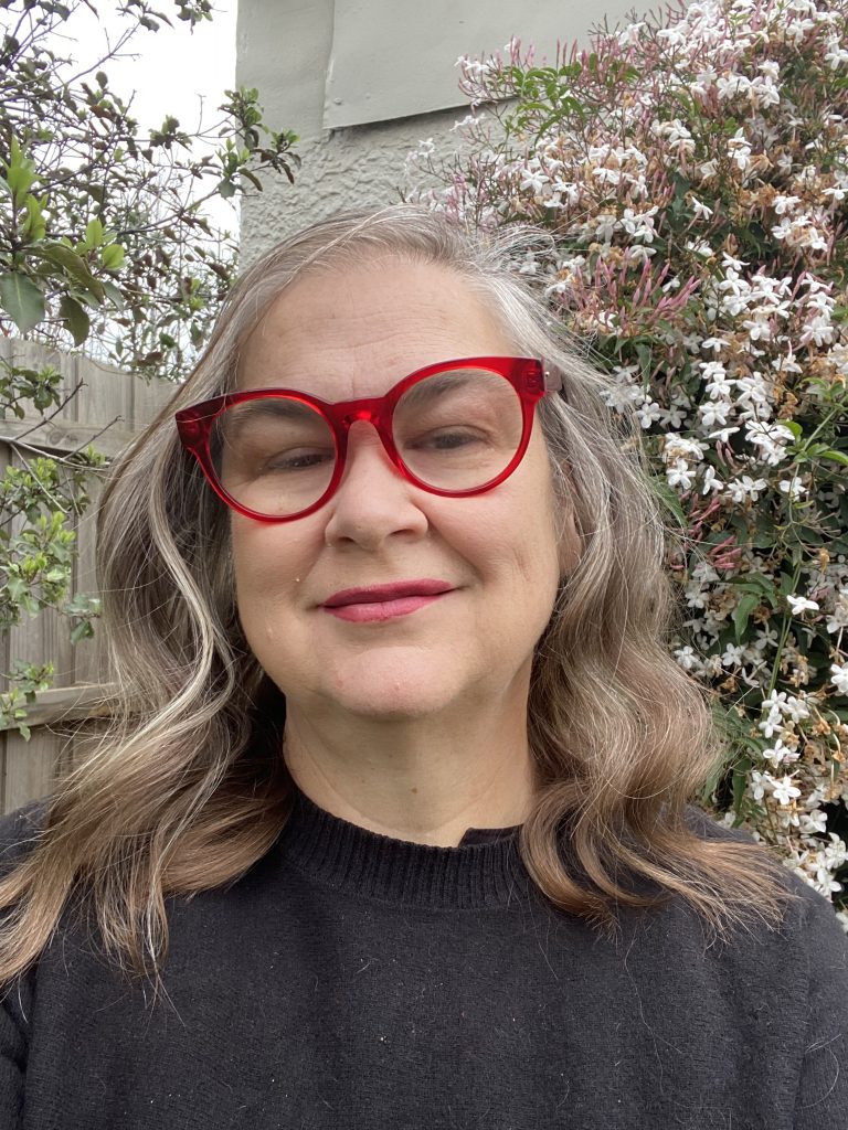 Keynote speaker Professor Cat Hope. Professor Hope is wearing red glasses, smiling and looking at the camera. Behind her is a paling fence and some climbing jasmine in bloom. 