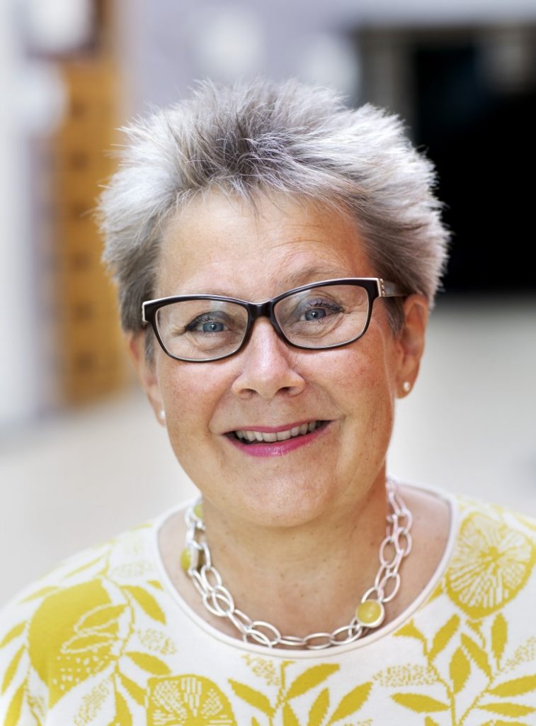 Keynote speaker, Pia Shekhter, international IAML President. Pia is smiling and looking at the camera. She wears black glasses, a silver necklace with yellow stones, and a white top with a yellow citrus print.